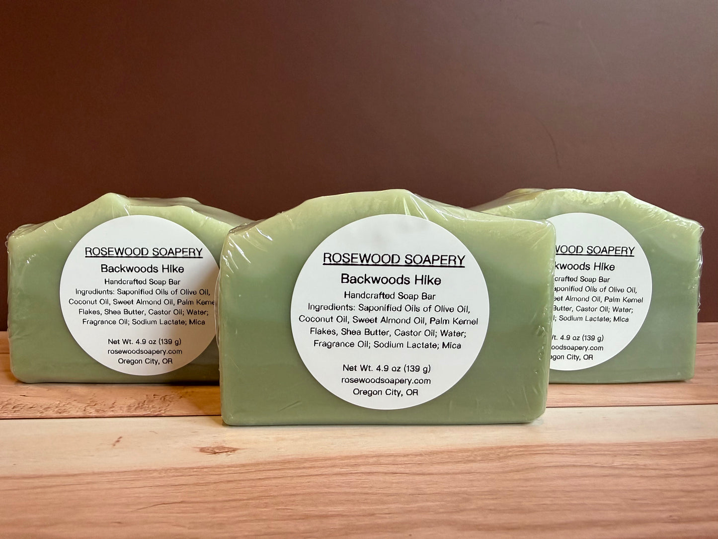 Three bars of Rosewood Soapery Backwoods Hike soap on a wooden surface with a brown background.
