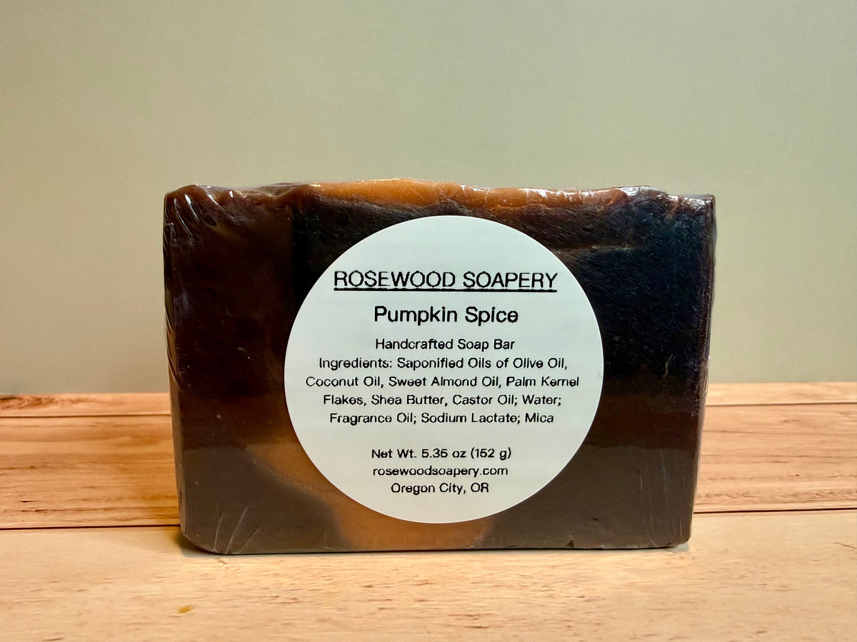 A rectangular bar of Rosewood Soapery Pumpkin Spice soap wrapped in clear plastic, featuring a white circular label, set on a wooden surface against a beige background.
