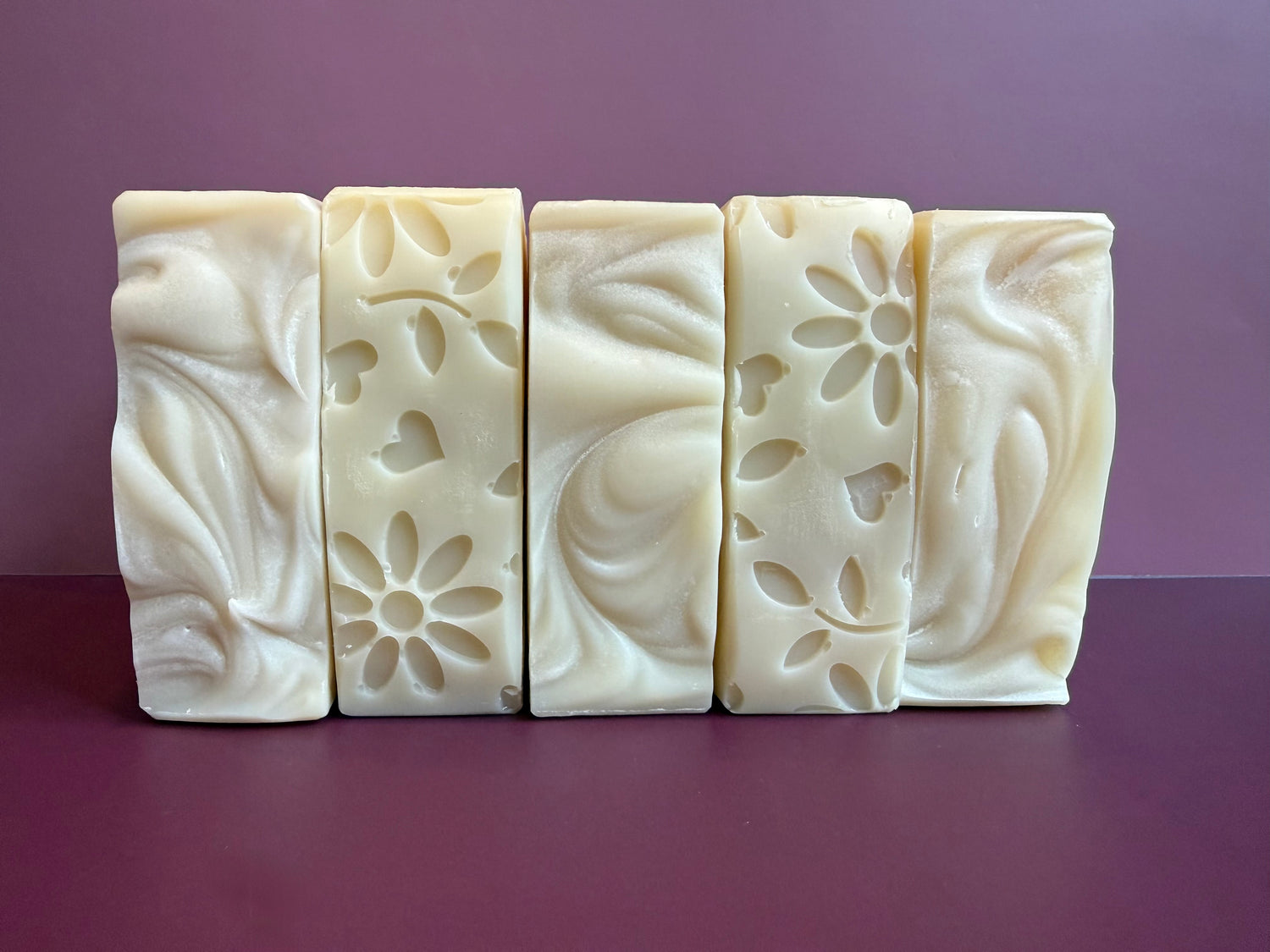 Five soaps lined up in a row, alternating between mounded tops and floral bottoms, against a purple background. 