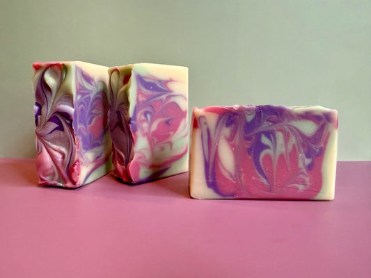 Three handmade marbled soap bars with swirls of pink, purple, and cream, displayed on a pink surface against a light gray background.