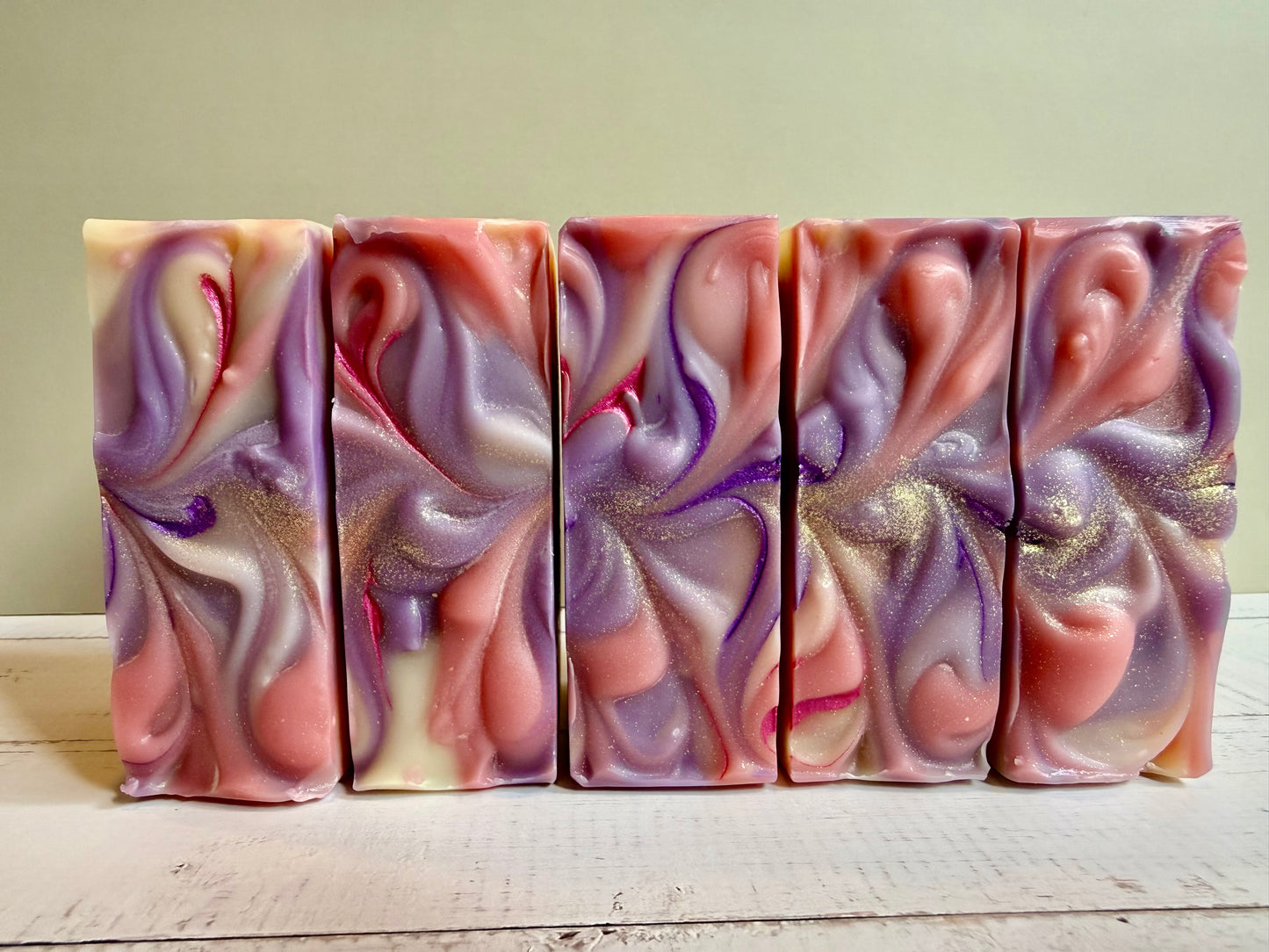 Five bars of swirled soap with pink, purple, and gold colors on a wooden surface.