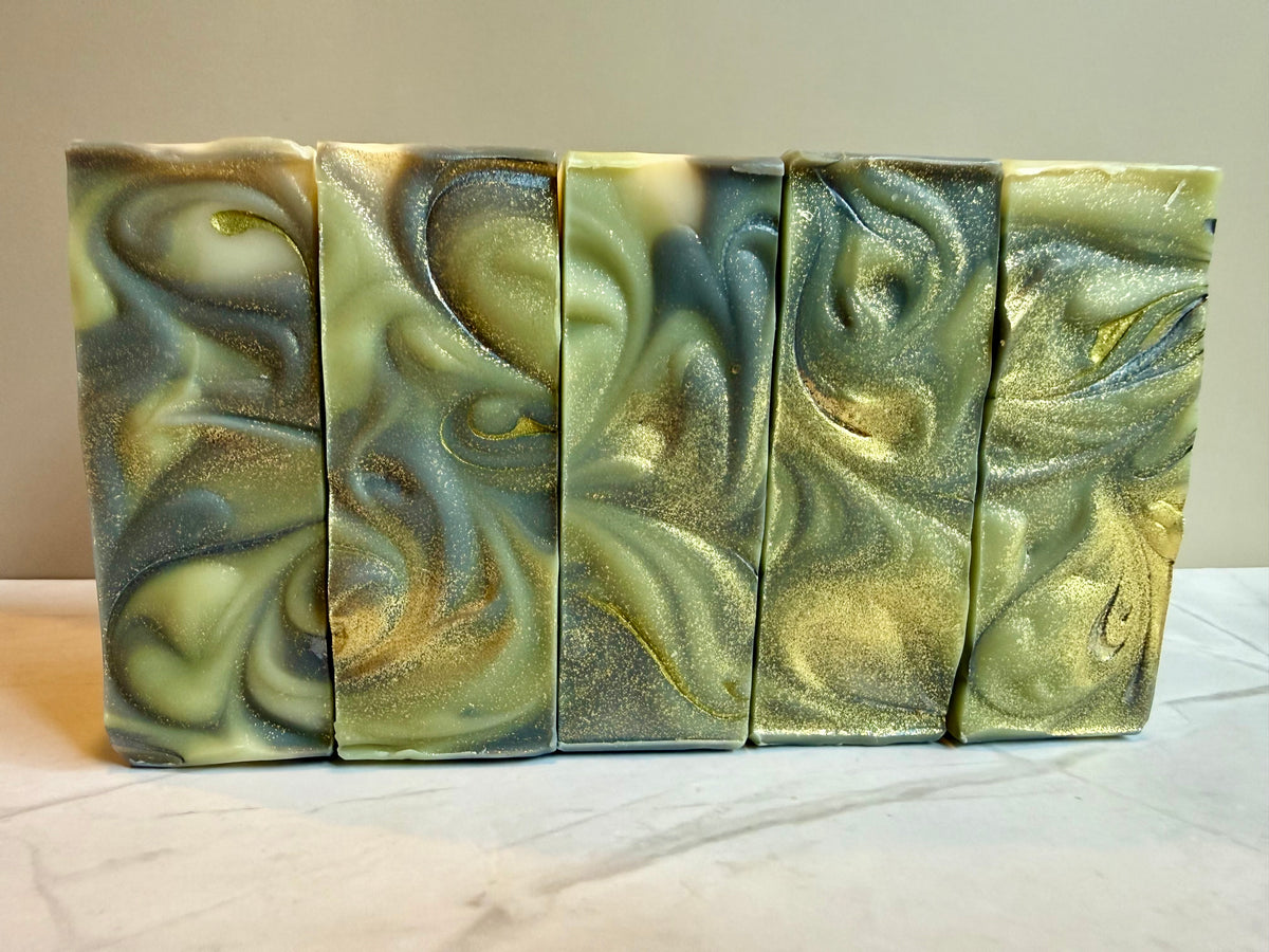 Five rectangular bars of handmade soap with a marbled swirl pattern in olive green, slate blue, and gold glitter, arranged side-by-side on a light marble surface against a neutral beige background.
