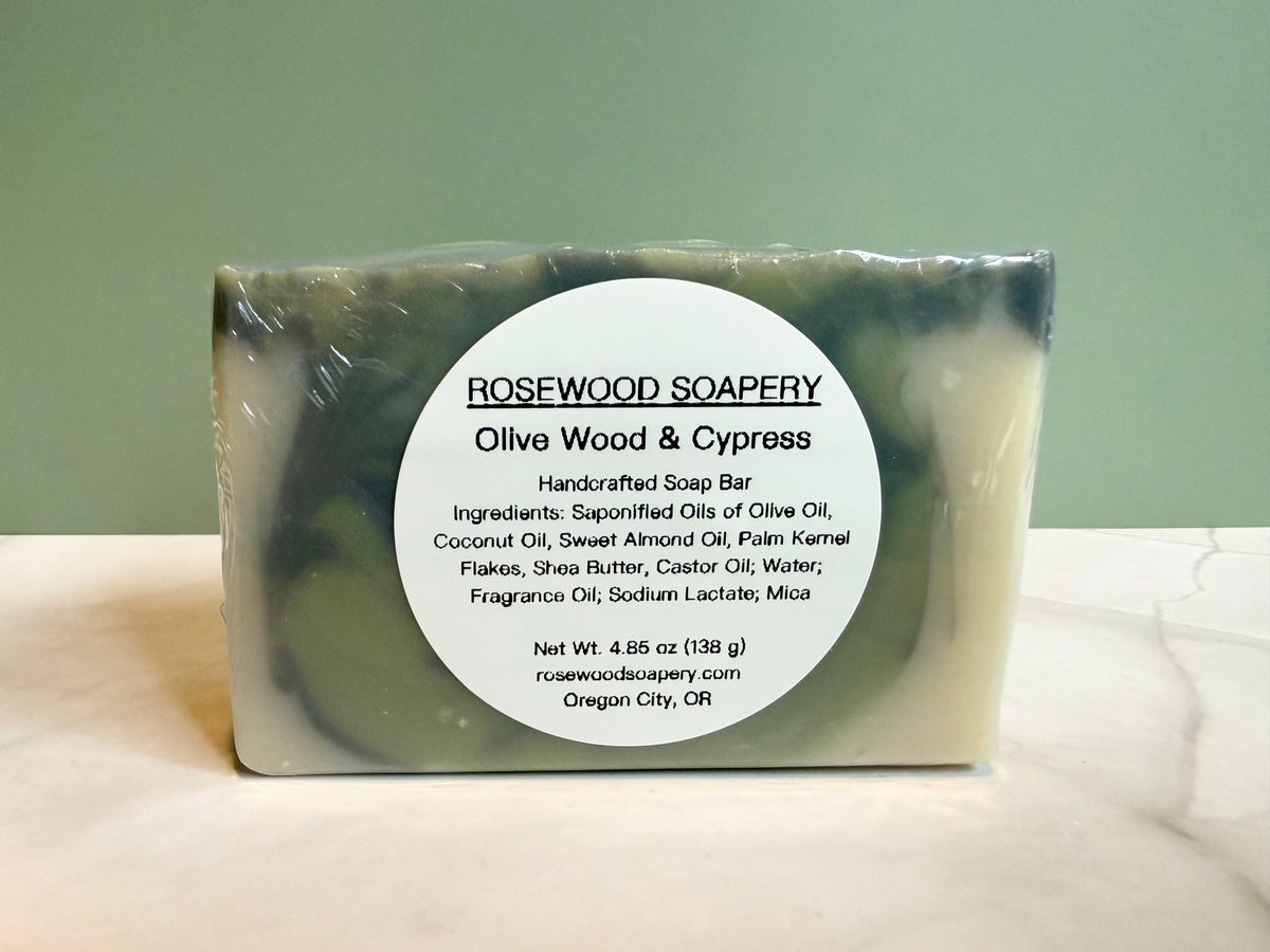 A rectangular bar of soap wrapped in clear plastic, featuring a white circular label that reads 'ROSEWOOD SOAPERY Olive Wood & Cypress.' The soap has a marbled color identity of olive green and pale cream.