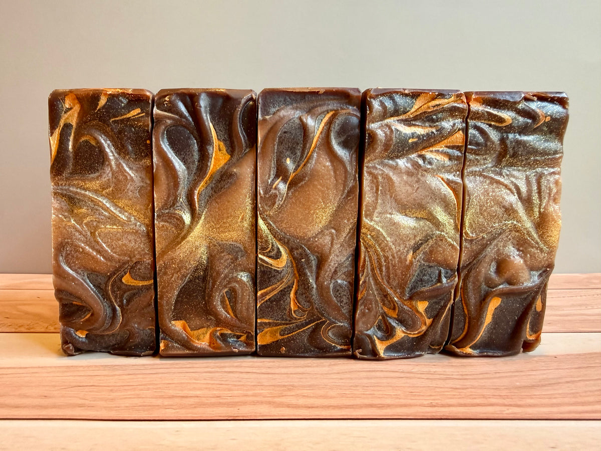 Four rectangular bars of handmade soap with a dark brown, orange and gold marbled swirl pattern, arranged side-by-side on a light wood surface against a neutral beige background.