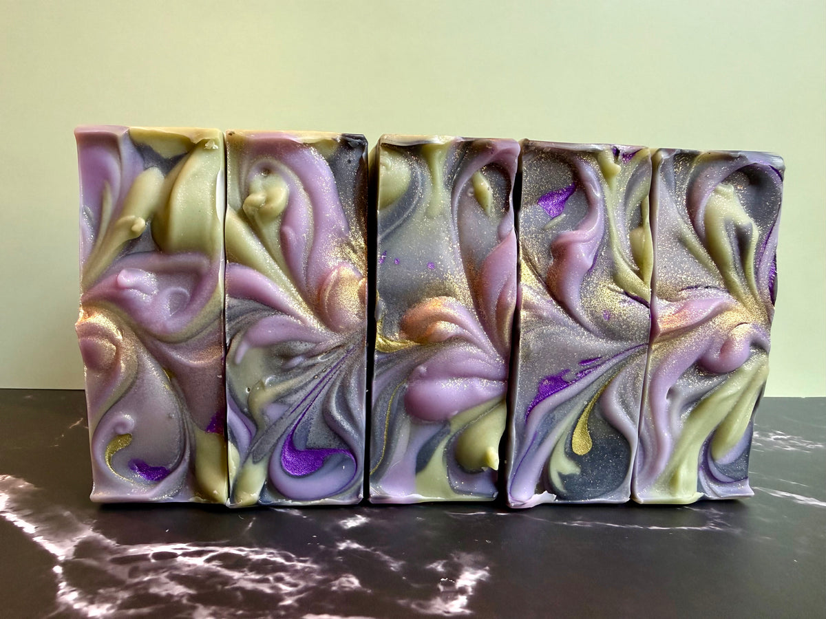 Five rectangular bars of marbled artisan soap with purple, lavender, olive green, and gold swirls, arranged side-by-side on a dark marble surface against a pale green background.