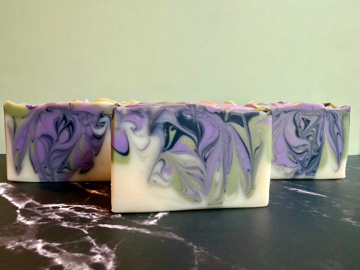 Three rectangular handmade soap bars with purple, lavender, and white marbled swirls, displayed on a dark marble surface against a pale green background.
