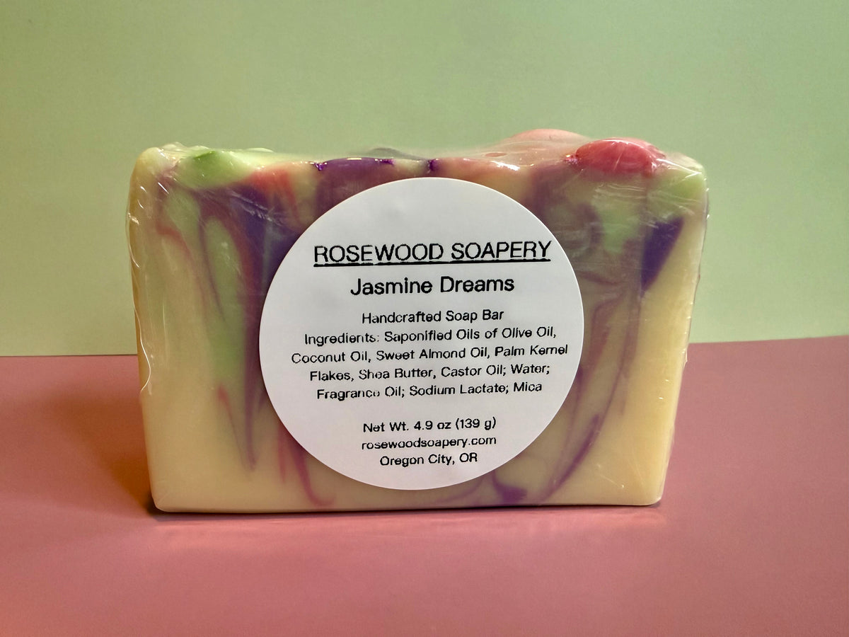 A rectangular handmade soap bar wrapped in clear plastic with a large white circular label reading 'ROSEWOOD SOAPERY Jasmine Dreams.' The soap has a creamy off-white base with marbled swirls of purple, pink, and green.