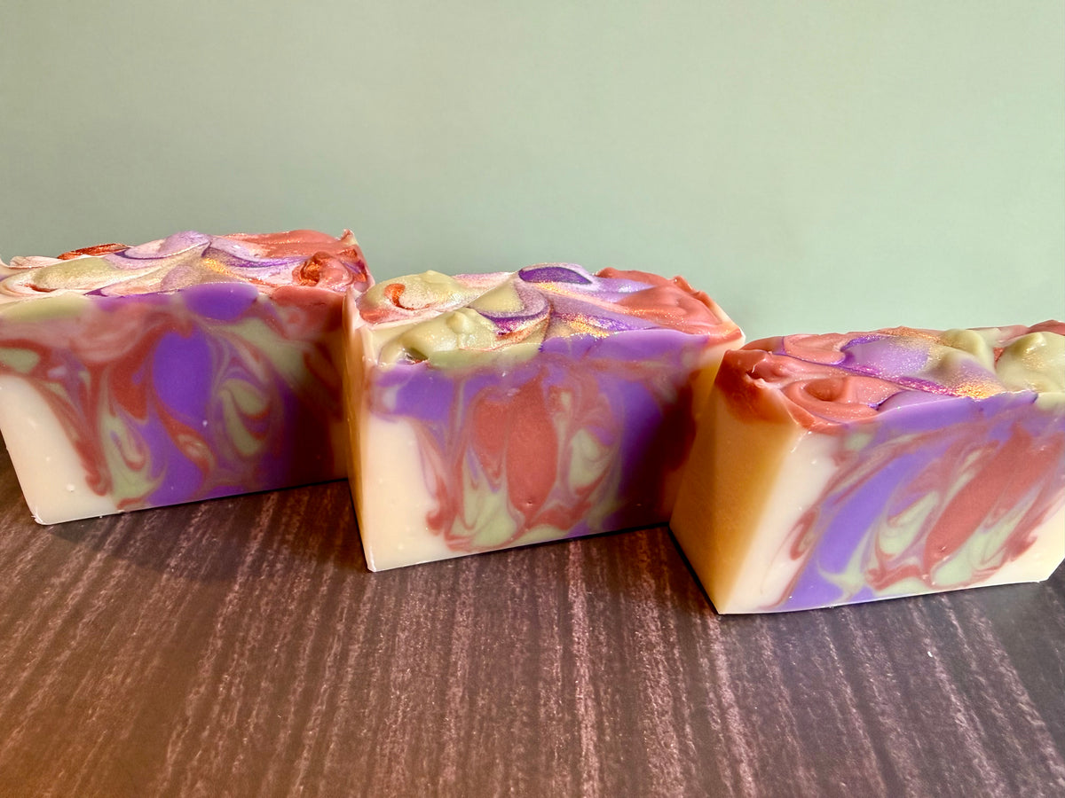 Three handmade rectangular bars of soap with marbled swirls of purple, brown, cream, and light green, displayed on a dark wood surface against a pale green background.