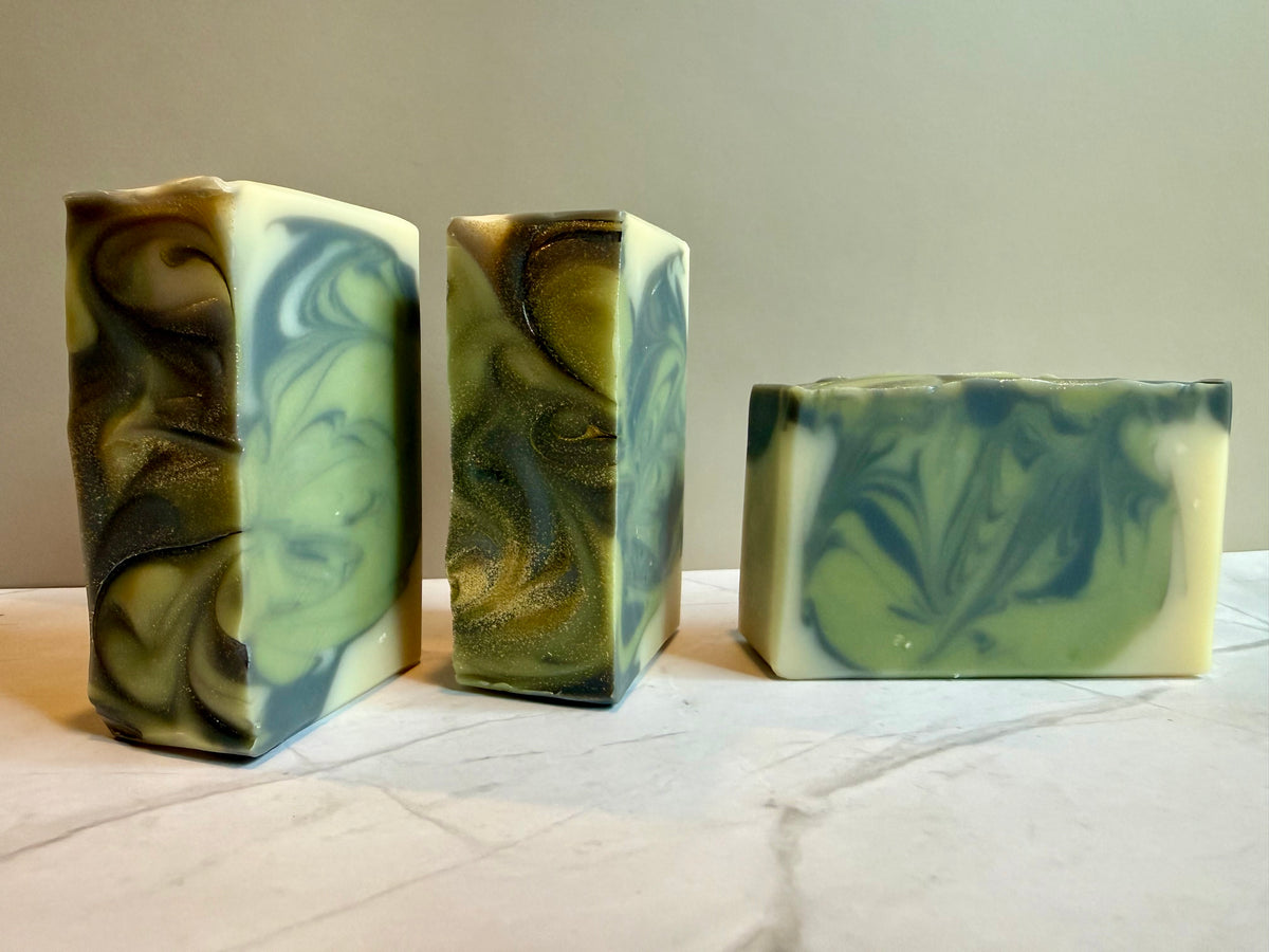 Three handmade marbled soap bars in green, cream, and dark gray swirls, displayed on a light marble surface against a neutral beige background.