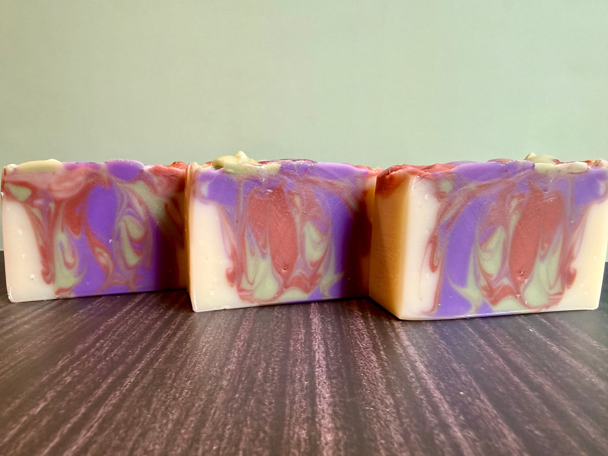 Three rectangular handmade soap bars with a marbled swirl pattern of purple, brown, cream, and light green, displayed on a dark wood surface against a pale green background.
