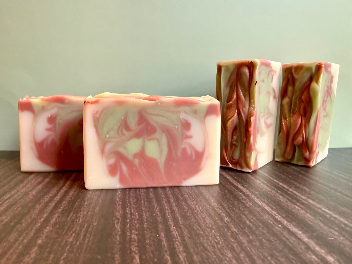 Four handmade rectangular bars of soap with a marbled swirl pattern of brown, sage green, and off-white, arranged on a dark wood surface against a pale green background.