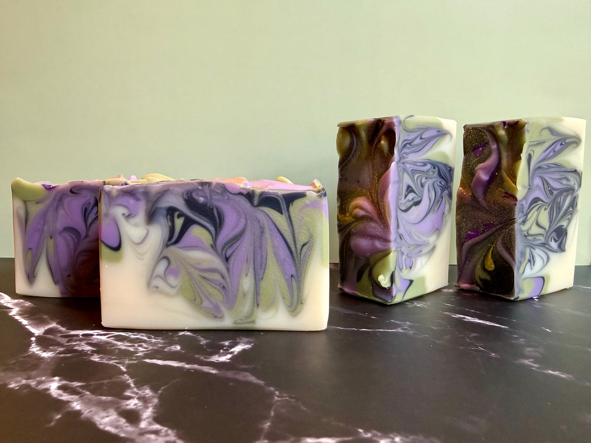 Four handmade marbled soaps with purple, white, and olive green swirls, arranged on a dark marble surface against a light green wall.