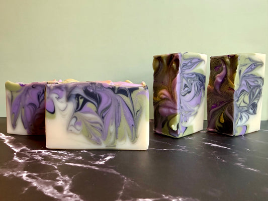 Four handmade marbled soaps with purple, white, and olive green swirls, arranged on a dark marble surface against a light green wall.