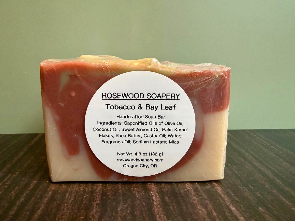 A rectangular bar of Rosewood Soapery Tobacco & Bay Leaf handcrafted soap bar, wrapped in clear plastic and featuring a white circular label, set on a dark wooden surface against a soft green background.