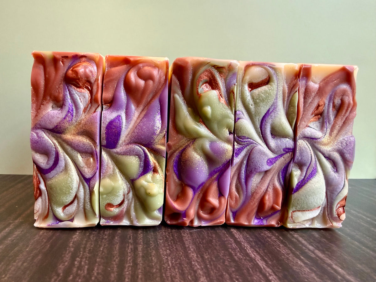 Five handmade marbled soap bars with swirls of purple, brown, and pale green, standing upright on a dark wooden surface against a light beige wall.