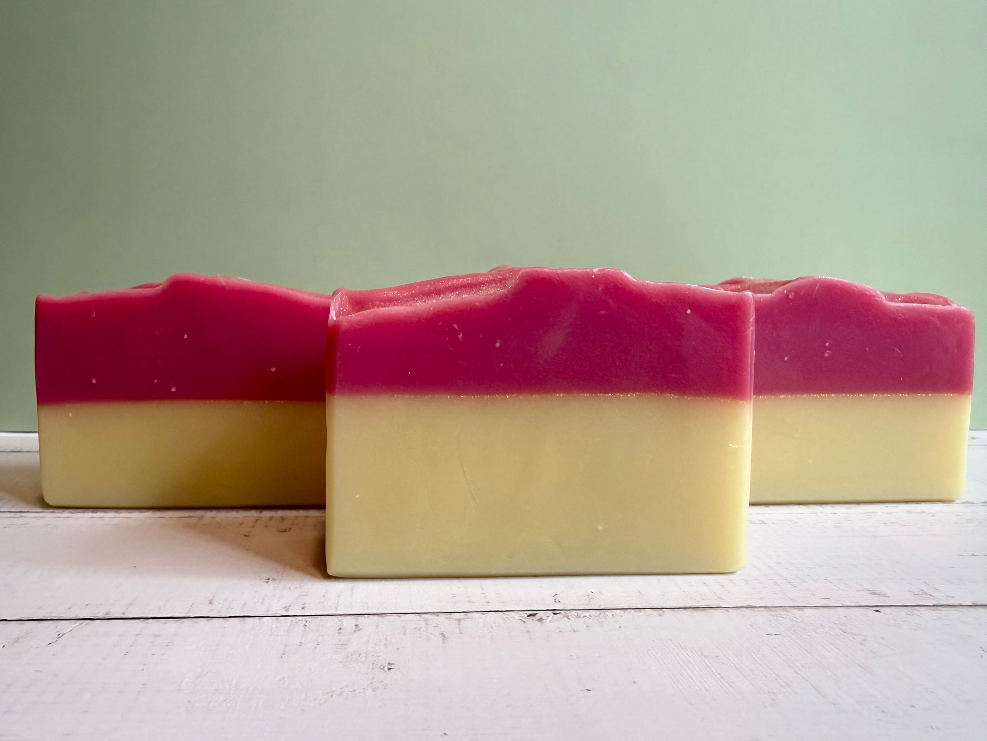 Three bars of soap with a pink and yellow layers on a wooden surface against a green wall. 