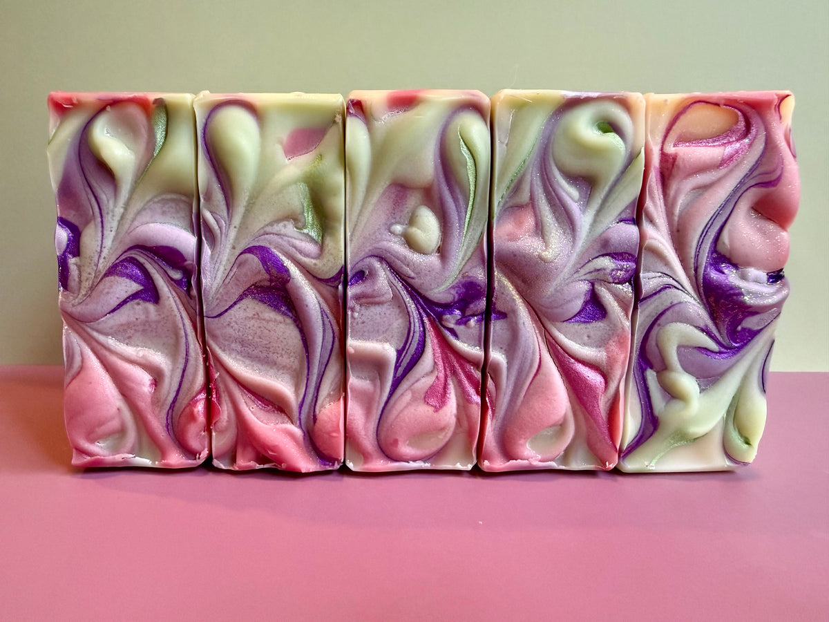 Five rectangular bars of marbled artisan soap with pink, purple, and pale green swirls, arranged side-by-side on a pink surface against a light beige background.