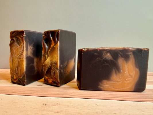 Three rectangular handmade soap bars with a marbled swirl pattern of dark brown and orange, displayed on a light wooden surface against a neutral gray background.