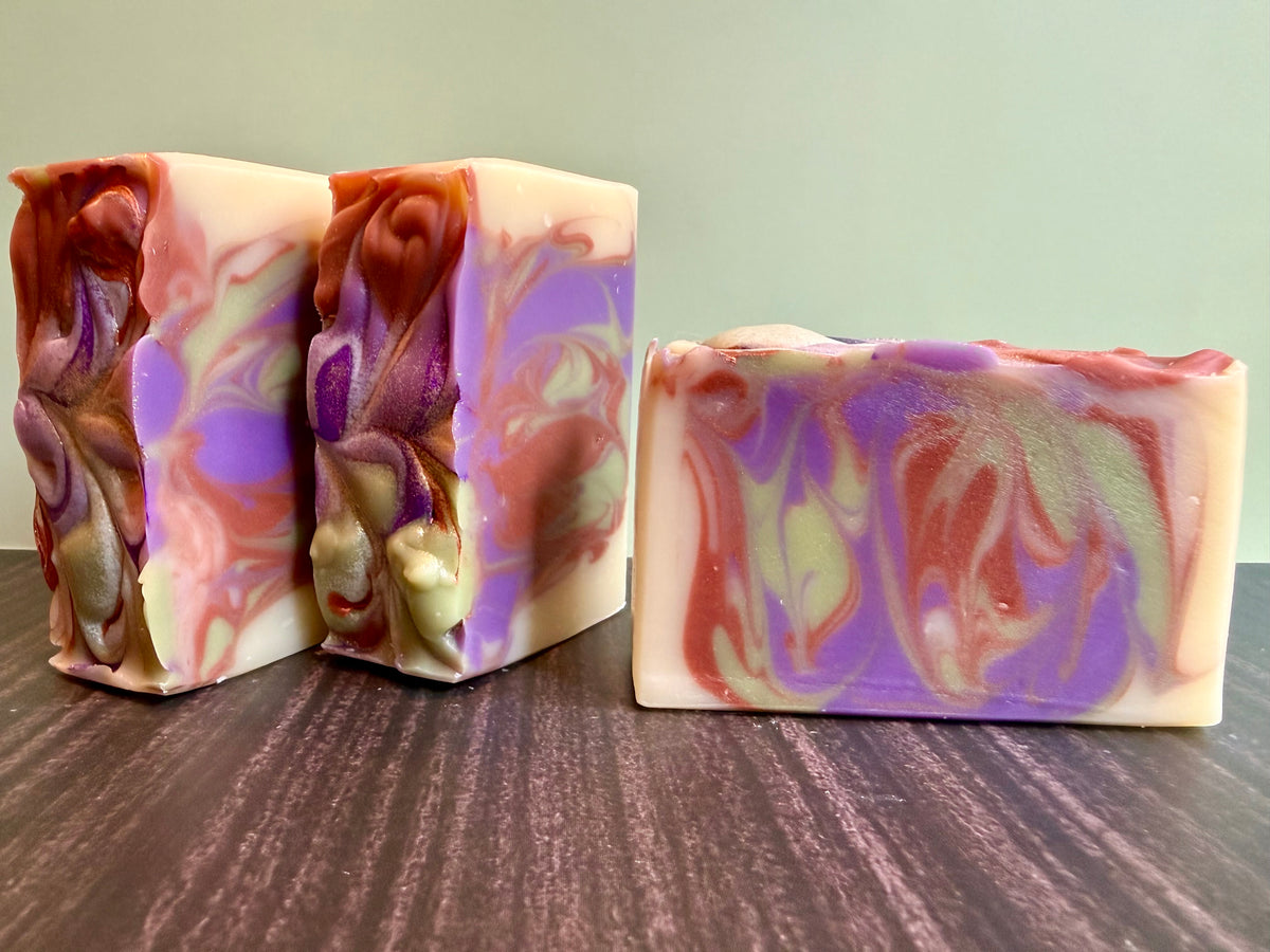 Three handmade marbled soap bars with swirls of purple, brown, green and cream, displayed on a dark wood surface against a light green background.