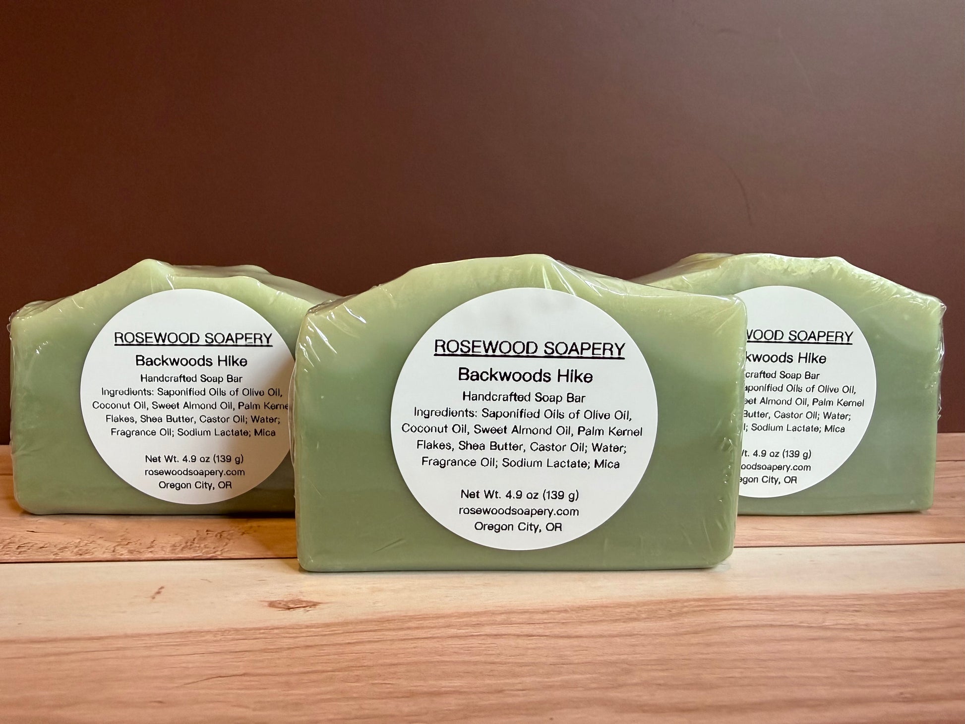 Three bars of Rosewood Soapery Backwoods Hike soap on a wooden surface with a brown background.