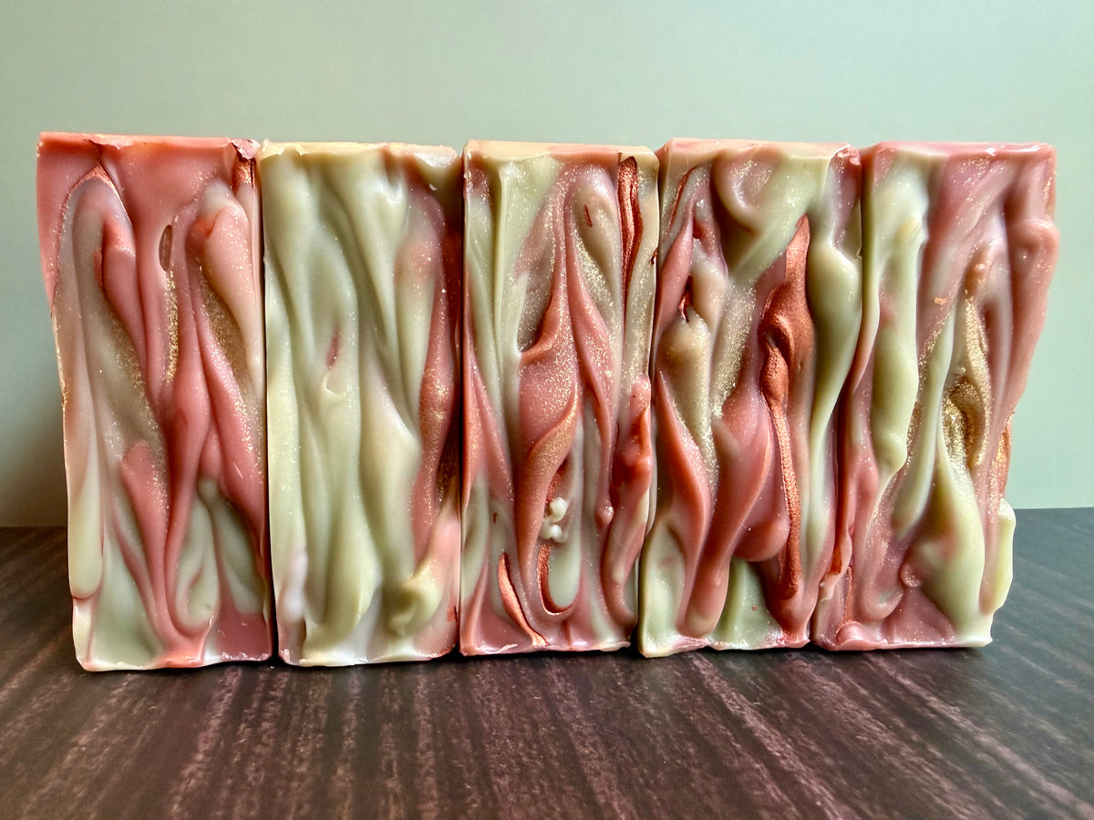 Four rectangular bars of marbled handmade soap with brown, cream, and pale green swirls, standing upright on a dark wooden surface against a light green wall.