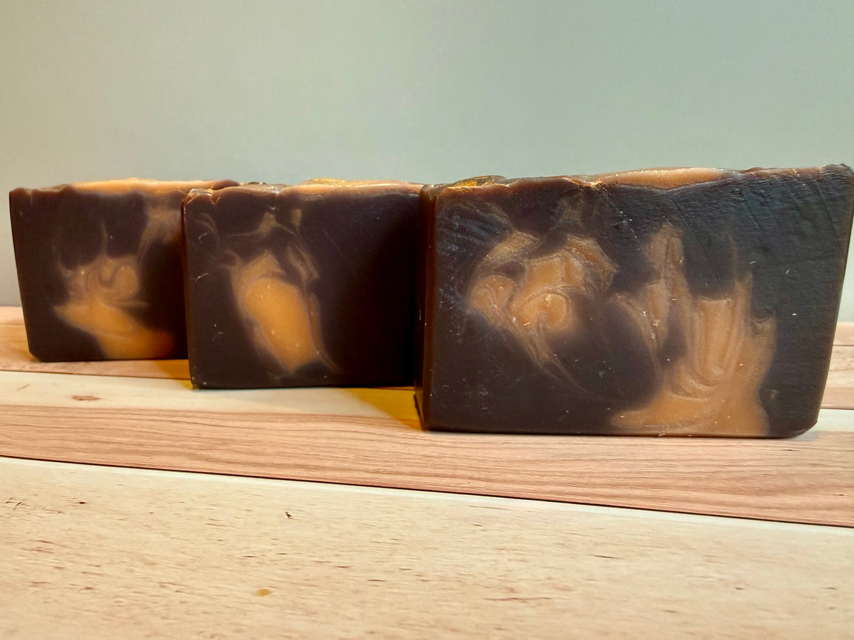Three rectangular bars of handmade soap with a dark brown and orange swirl pattern, displayed on a light wooden surface against a soft gray background.