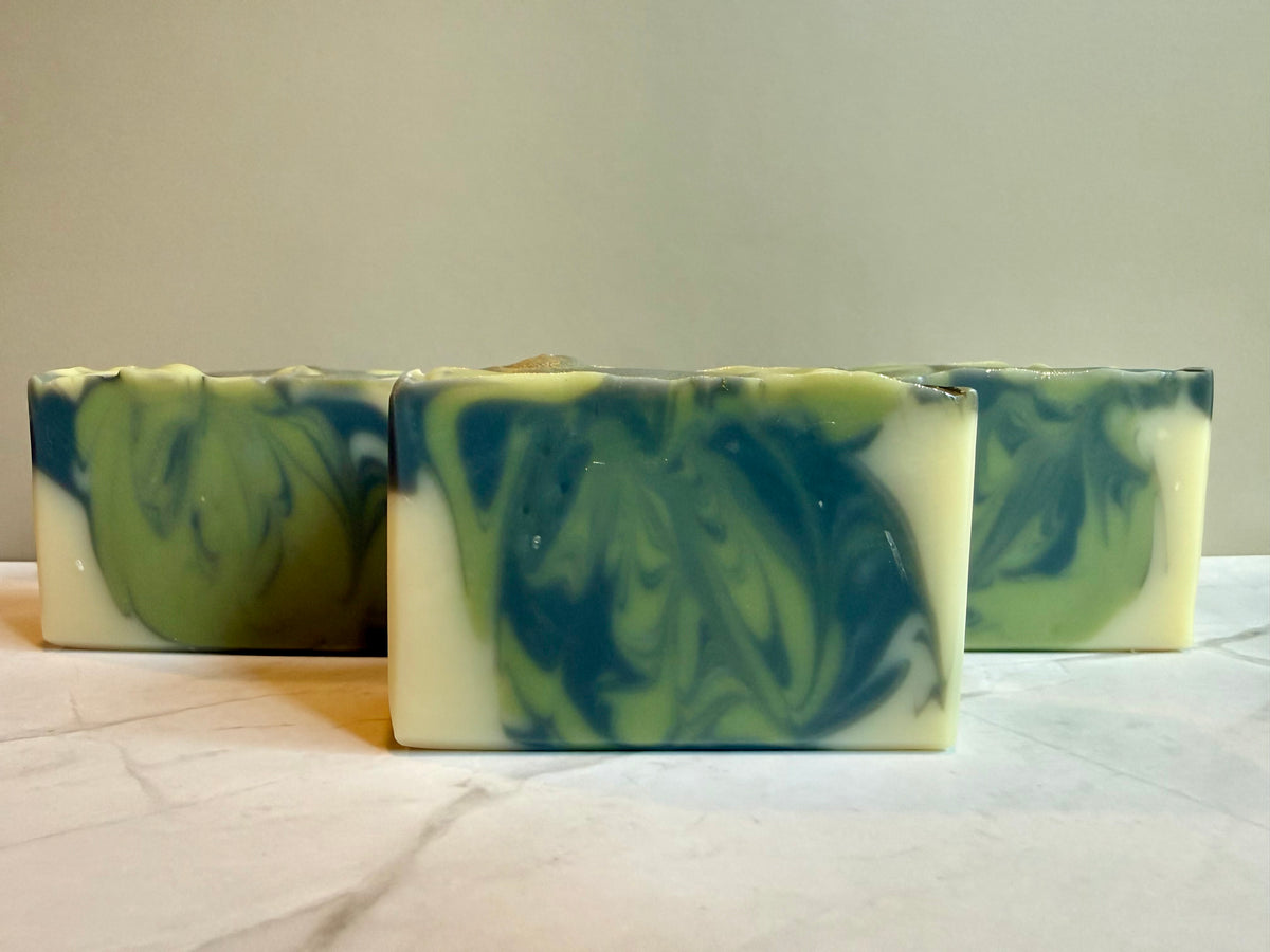 Three rectangular handmade soap bars with a green and dark blue marbled swirl pattern, arranged on a light marble surface against a neutral beige background.