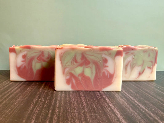Three rectangular handmade soap bars with brown and light green marbled swirls, arranged on a dark wood surface against a pale green background.