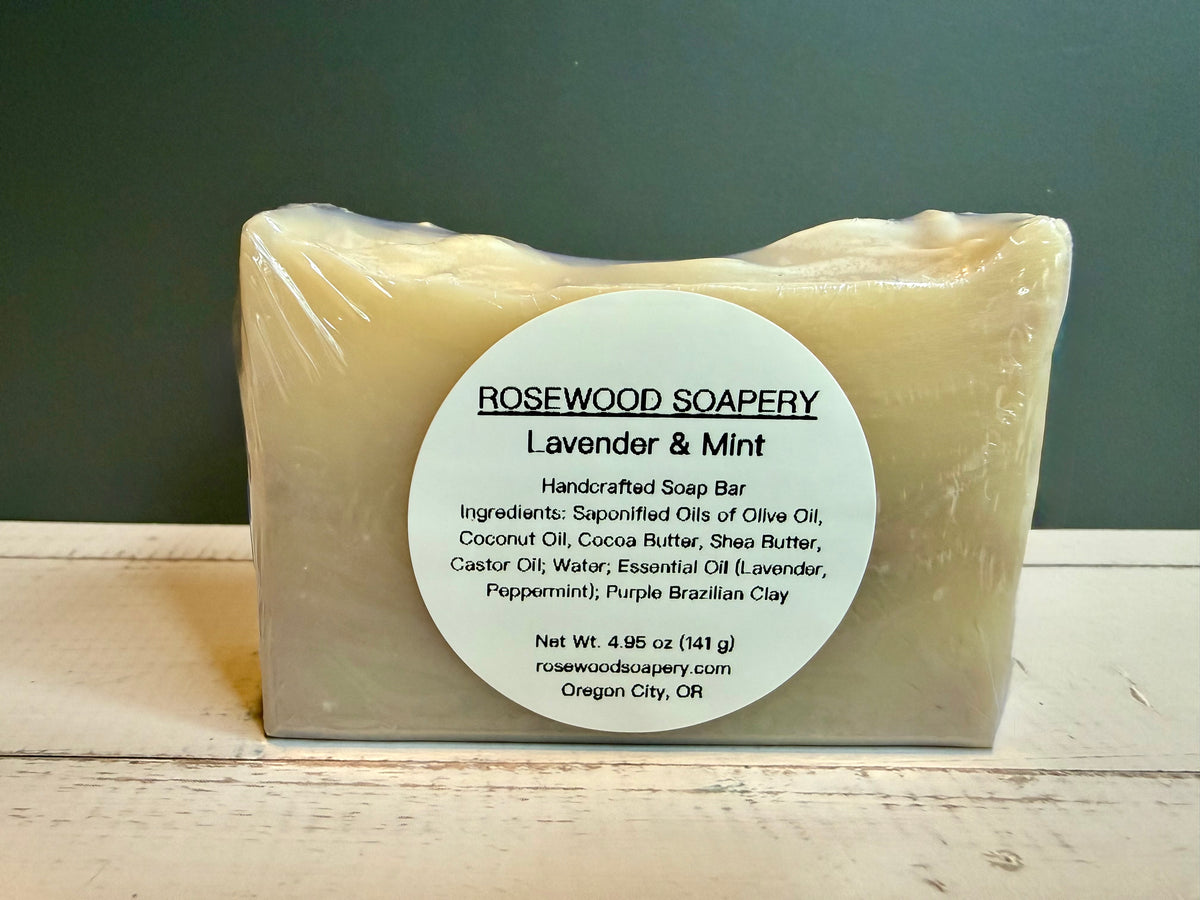 A rectangular bar of Rosewood Soapery Lavender & Mint soap wrapped in clear plastic, featuring a large white circular label, set against a dark green background and light wood surface.