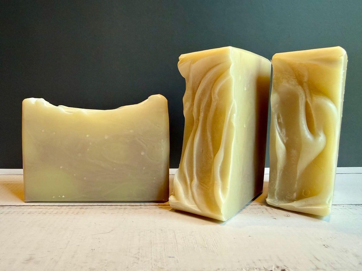 Three handmade soap bars in natural pale yellow and cream tones, displayed on a light wooden surface against a dark gray background.