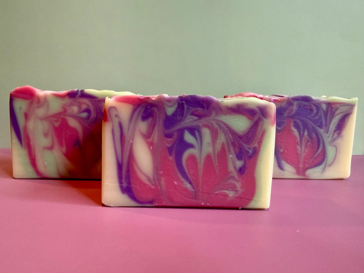 Three rectangular handmade soap bars with a marbled swirl pattern of pink, purple, and white, arranged on a pink surface against a light green background.