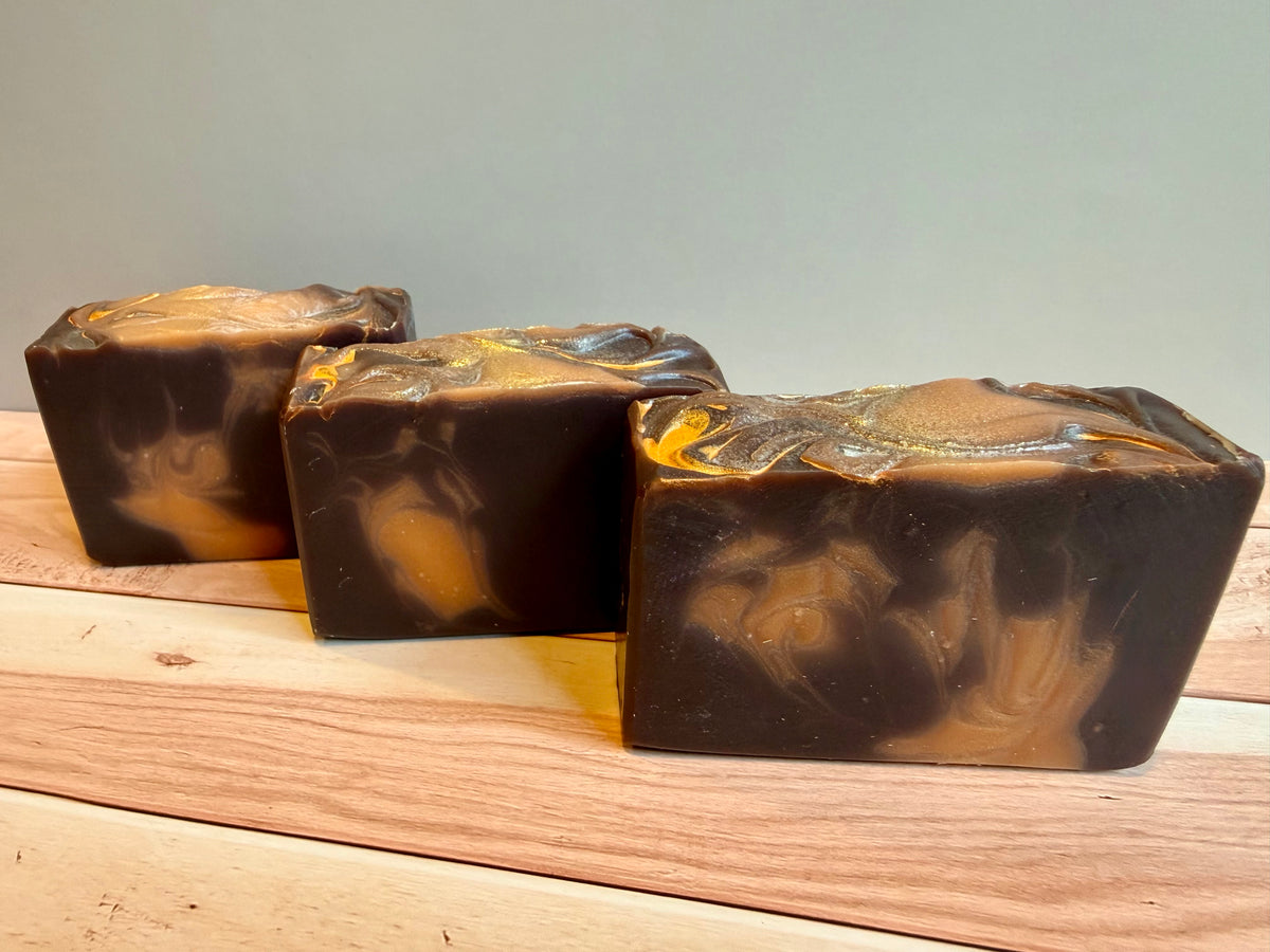 Three rectangular bars of handmade soap with a dark brown and orange marbled swirl pattern, arranged on a light wood surface against a neutral gray background.