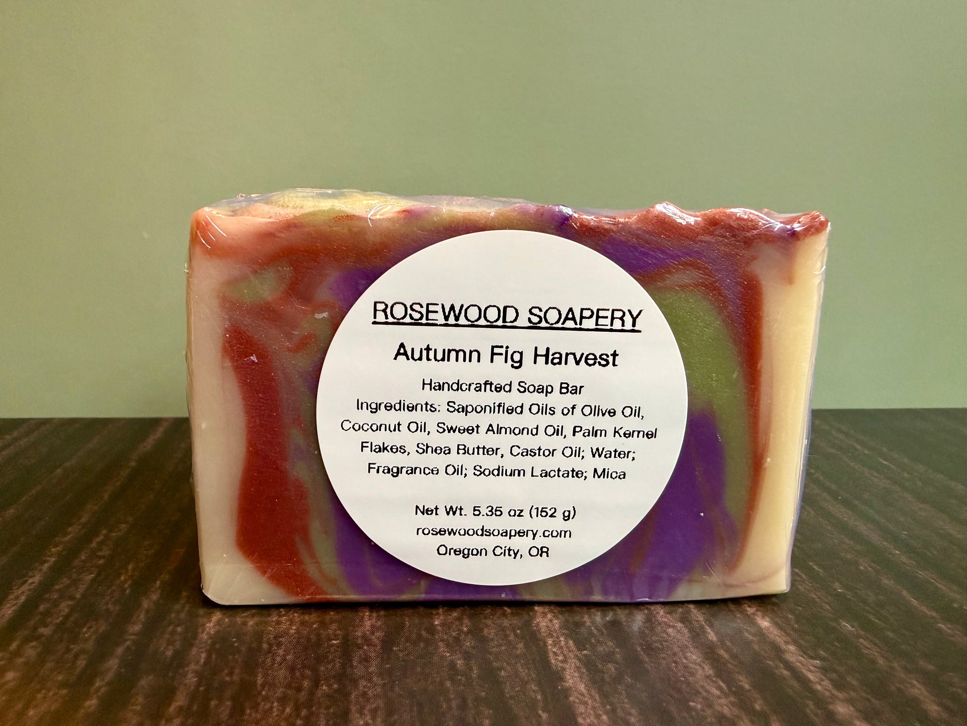 A rectangular bar of handmade soap from Rosewood Soapery, labeled 'Autumn Fig Harvest,' standing upright on a dark wooden surface against a light green background. The soap has marbled swirls of cream, brown, purple, and green, with a large white circular label on the front.