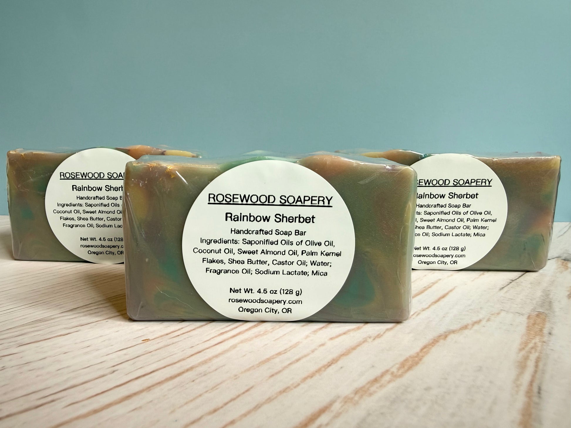 Three bars of Rosewood Soapery Rainbow Sherbet soap on a wooden surface with a light blue background.