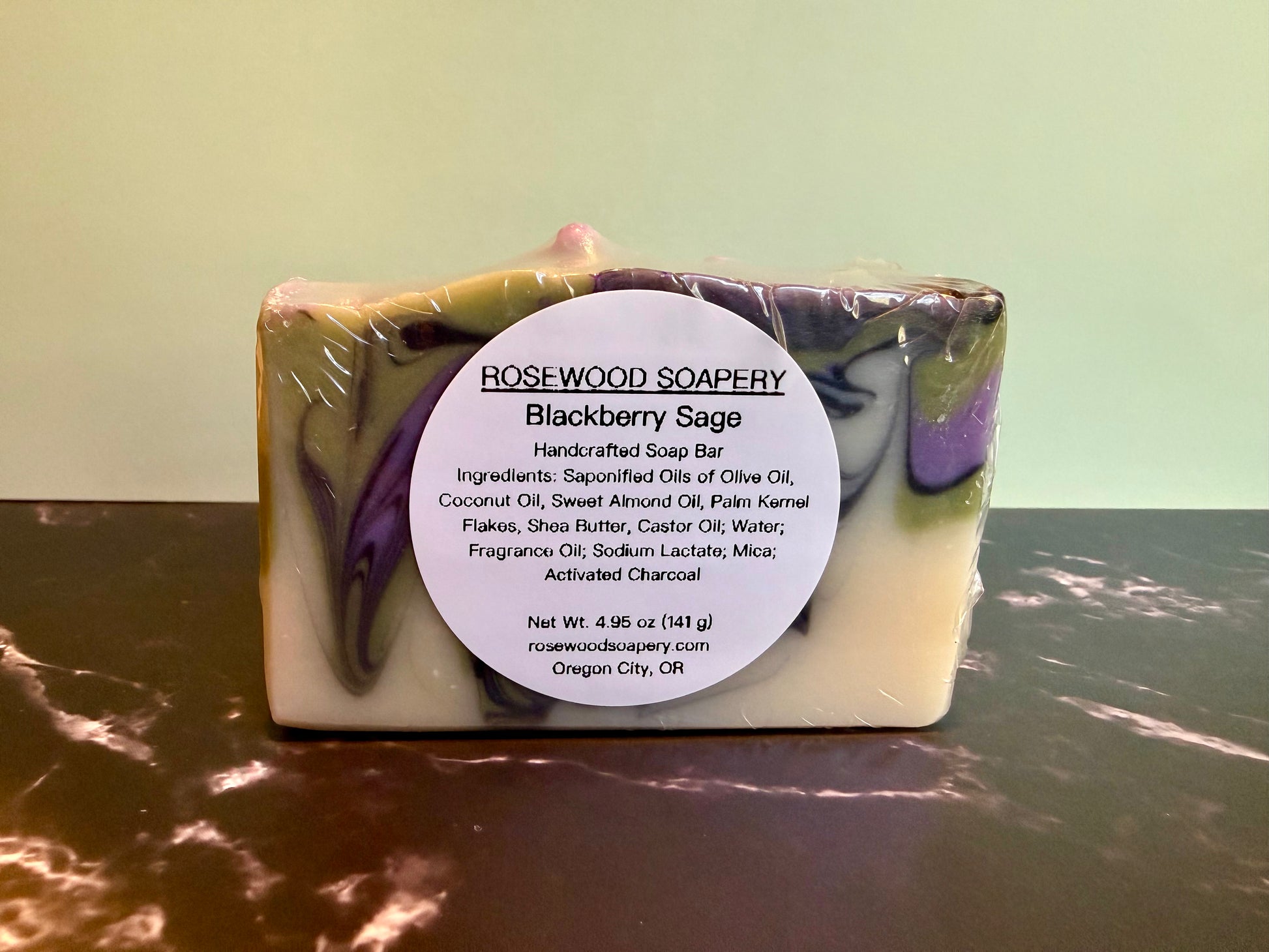 A rectangular handmade soap bar wrapped in clear plastic with a large white circular label reading 'ROSEWOOD SOAPERY Blackberry Sage.' The soap has a marbled color identity with white, purple, and olive-green tones.