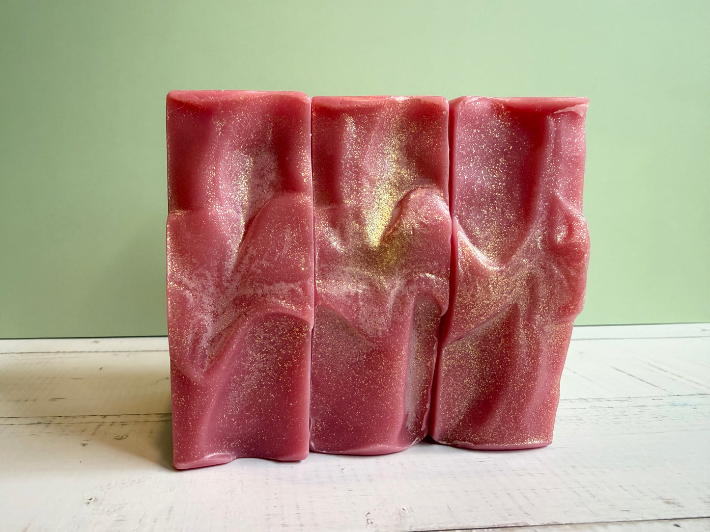 Three pink soap bars with textured tops on a light wooden surface against a green wall.