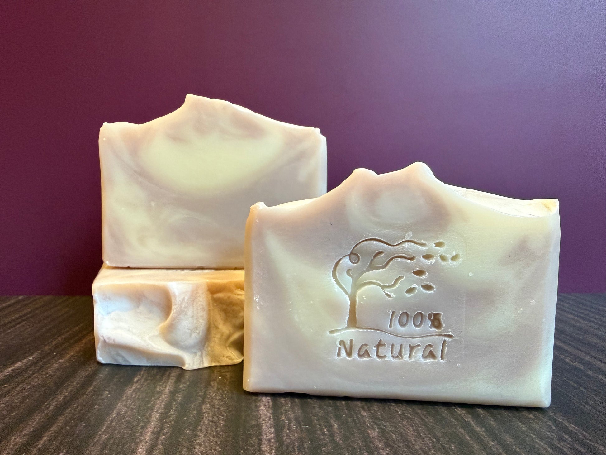 Three bars of soap with a '100% Natural' logo on a wooden surface against a purple background