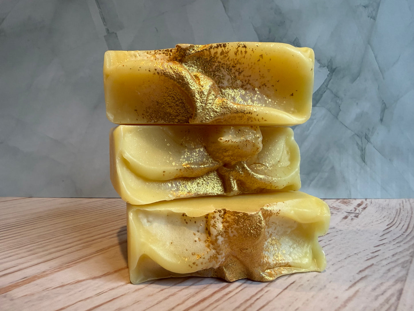 Three bars of yellow soap with gold flakes stacked on a wooden surface with a marble background