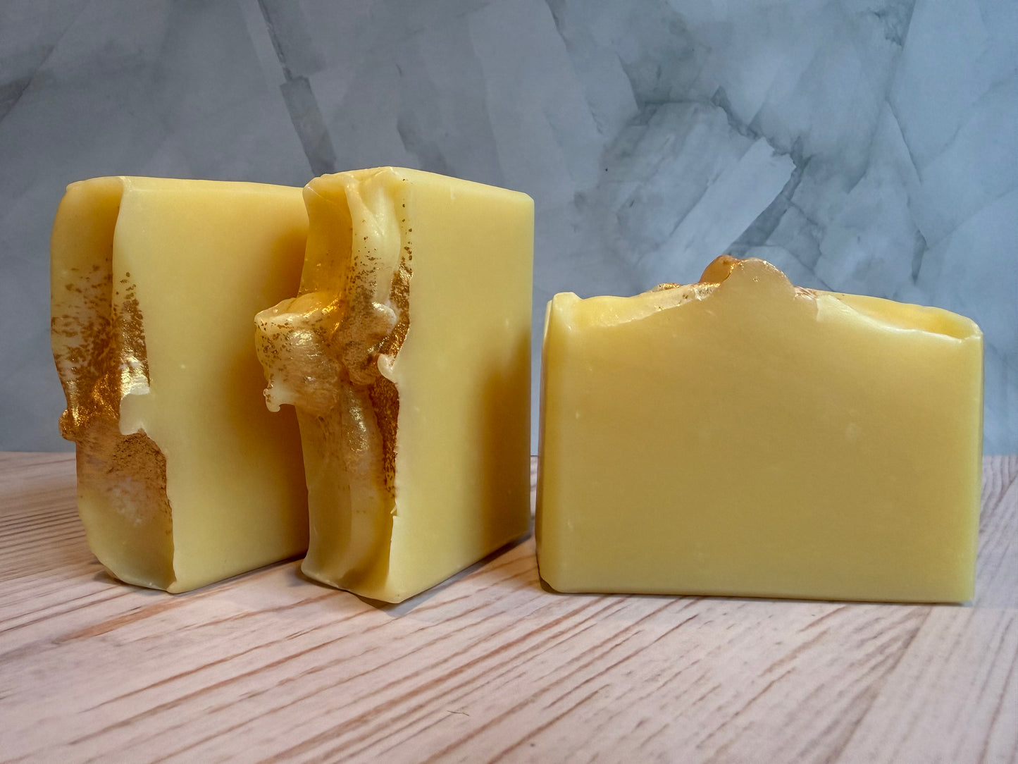 Three pieces of yellow soap on a wooden surface with a marble-patterned wall in the background.